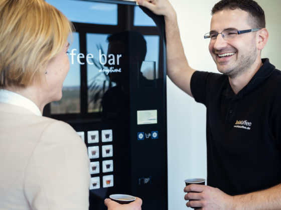 eine Frau und ein Mann stehen lachend vor einem Kaffee- und Heissgetränke Standgerät